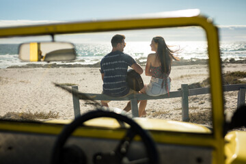 Happy caucasian couple next to beach buggy by the sea playing guitar