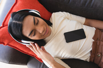 Smiling mixed race gender fluid man lying on couch wearing wireless headphones using smartphone
