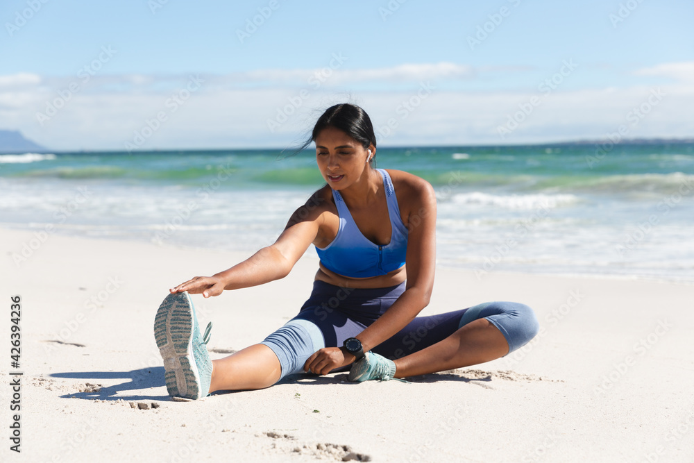 Wall mural mixed race woman exercising on beach wearing wireless earphones sitting and stretching