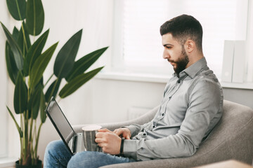 Handsome young man of oriental appearance works at home, sitting in a chair with a laptop