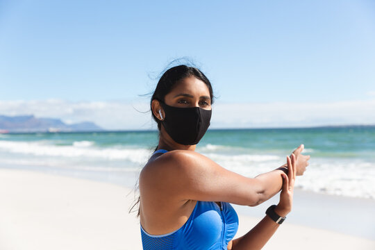 Mixed race woman exercising on beach wearing face mask and wireless earphones stretching arms - Powered by Adobe