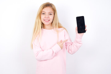 Excited beautiful caucasian little girl wearing pink hoodie over white background holding and pointing with finger at smartphone with blank screen. Advertisement concept.