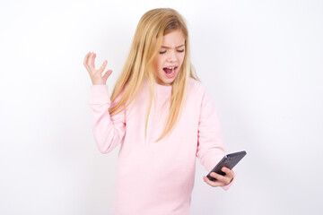 Photo of outraged annoyed beautiful caucasian little girl wearing pink hoodie over white background holds cell phone, makes call, argues with colleague,  expresses negative emotions. People and anger.