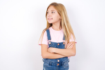 Charming thoughtful beautiful Caucasian little girl wearing denim overalls over white background stands with arms folded concentrated somewhere with pensive expression thinks what to do