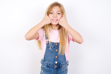 Happy beautiful Caucasian little girl wearing denim overalls over white background  keeps fists on cheeks smiles broadly and has positive expression being in good mood