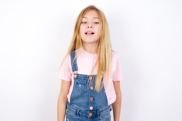 Funny beautiful Caucasian little girl wearing denim overalls over white background makes grimace and crosses eyes plays fool has fun alone sticks out tongue.