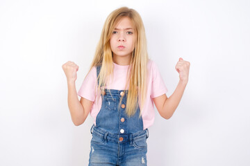 Irritated beautiful Caucasian little girl wearing denim overalls over white background blows cheeks with anger and raises clenched fists expresses rage and aggressive emotions. Furious model