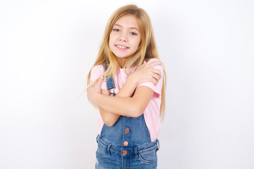 Charming pleased beautiful Caucasian little girl wearing denim overalls over white background embraces own body, pleasantly feels comfortable poses. Tenderness and self esteem concept