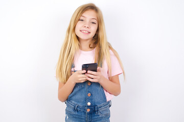 beautiful Caucasian little girl wearing denim overalls over white background Mock up copy space. Using mobile phone, typing sms message