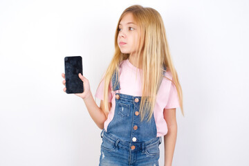 beautiful Caucasian little girl wearing denim overalls over blue background holds new mobile phone and looks mysterious aside shows blank display of modern cellular