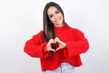 young beautiful brunette woman wearing red knitted sweater over white wall smiling in love showing heart symbol and shape with hands. Romantic concept.