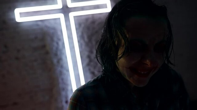 Portrait of a creepy clown on the background of a cross. The light blinks and illuminates the face in the makeup. Halloween party