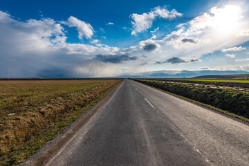 Empty asphalt road leading through green agricultural fields at spring, blue sky with white clouds.