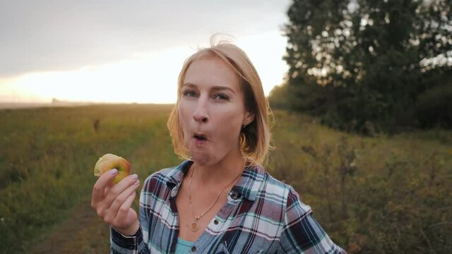 Happy blonde woman eating green apple at morning in the countryside. Close up of smiling woman eat apple fruit outdoor. Portrait of young woman eating apple. Pretty girl eat apple. Female healthy diet