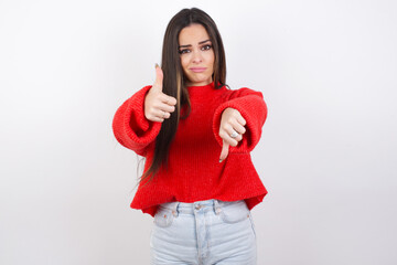 young beautiful brunette woman wearing red knitted sweater over white wall feeling unsure making...