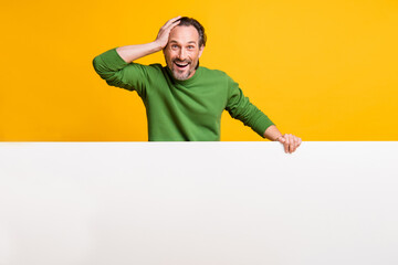 Photo of amazed man hold white banner palm head empty space wear green pullover isolated yellow color background