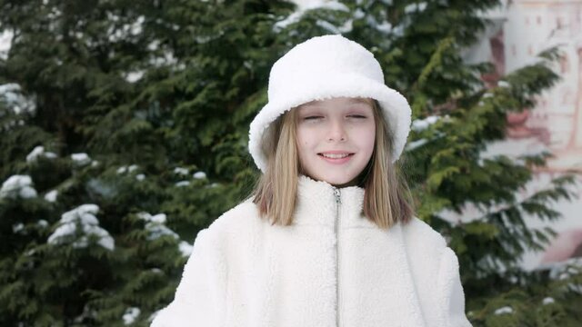 Portrait cute young woman in white hat and fur coat on evergreen thuja background on winter street. Smiling teenager girl looking camera on snowy thuja at winter outdoors