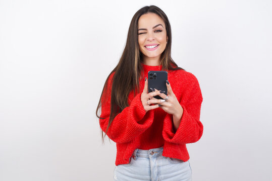 Pleased young beautiful brunette woman wearing red knitted sweater over white wall using self phone and looking and winking at the camera. Flirt and coquettish concept.