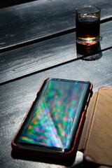 Vibrant rainbow rays reflect in mobile phone screen on black table with alcohol drink shot glass. Defocused multicolored beams and reflection in smartphone screen surface