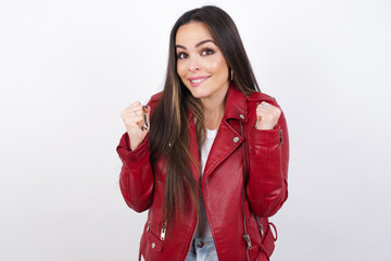 young beautiful brunette woman wearing red biker jacket over white wall clenches fists and awaits...