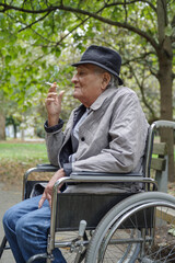 Obraz premium Disabled sad senior man smokes in outdoor while sitting in wheelchair