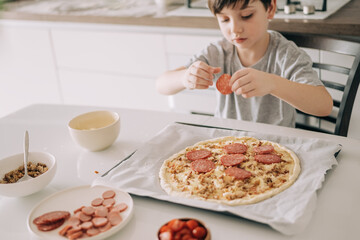 Little kid boy making pizza sitting at the table on the kitchen. Children helping in cooking lifestyle image