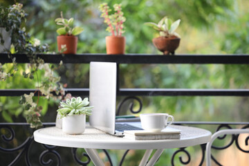 Silver laptop and coffee cup with notebooks on white table and plant pots on balcony outdoor