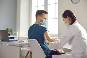Man in medical face mask getting flu shot during seasonal vaccination campaign at hospital or clinic. Female nurse or doctor giving modern Covid-19 vaccine to male patient during coronavirus outbreak
