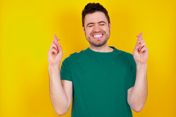 young Caucasian man wearing green T-shirt against yellow wall has big hope, crosses fingers,...