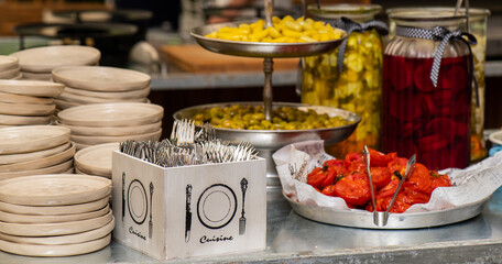 A table with box of forks ,jars and plates of pickles ,pile of plates