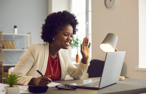 Friendly african american businesswoman holds an online meeting while sitting in the office. Woman waving hand greeting participants of a video call. Online business concept.