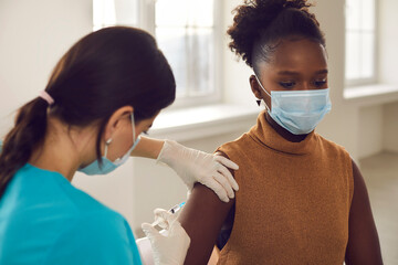 Young black african american woman patient in protective face mask sitting getting vaccination...