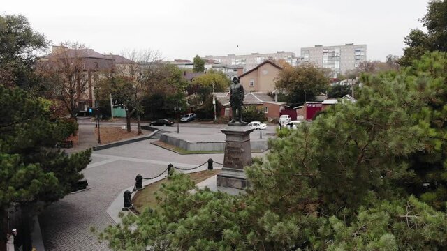 Shooting from a drone a male statue on a pedestal in a park among trees and a view of city blocks. statue of Peter the great in Taganrog
