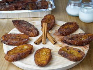 Traditional homemade and Delicious Spanish Torrijas making a circle and three cinnamon sticks in the middle on a round wooden plate decorated with its ingredients and a tray of torrijas