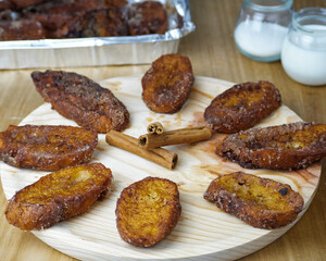 Traditional homemade and Delicious Spanish Torrijas making a circle and three cinnamon sticks in the middle on a round wooden plate decorated with its ingredients and a tray of torrijas
