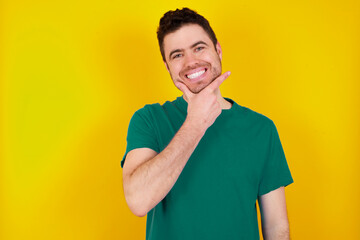 young Caucasian man wearing green T-shirt against yellow wall looking confident at the camera smiling with crossed arms and hand raised on chin. Thinking positive.