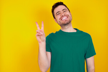 young Caucasian man wearing green T-shirt against yellow wall smiling with happy face winking at the camera doing victory sign. Number two.