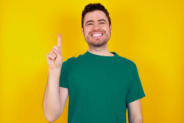 young Caucasian man wearing green T-shirt against yellow wall showing and pointing up with finger number one while smiling confident and happy.