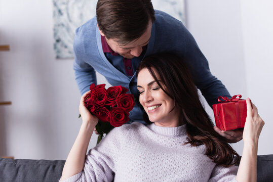 Man holding reses and gift box near wife on couch