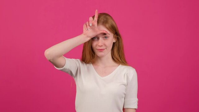 Young redhead woman with freckles doing loser gesture, pink background.