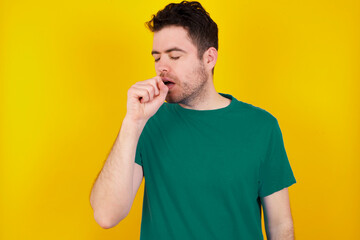 young Caucasian man wearing green T-shirt against yellow wall feeling unwell and coughing as symptom for cold or bronchitis. Healthcare concept.