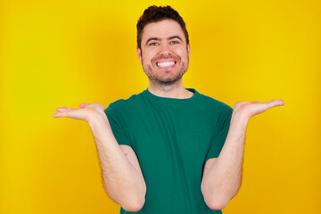 Cheerful cheery optimistic young Caucasian man wearing green T-shirt against yellow wall holding...