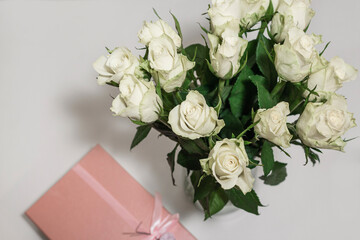 White tea roses stand in a glass vase, which is tied with a red ribbon and stands on the table. There is a gift nearby. in a pink box. Gift bouquet