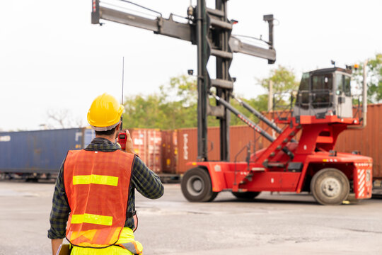 Engineer standing in front of Reach stacker vehicle in shipping yard. Inspector, dock worker using radio to command cargo forklift handling container. Logistics and transportation concepts.
