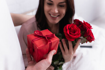 Man holding gift box near cheerful wife with flowers on blurred background on bed