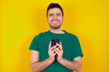 young handsome caucasian man wearing green t-shirt against yellow background enjoys distant communication, uses mobile phone, surfs fast unlimited internet, has pleasant smile, makes shopping online,