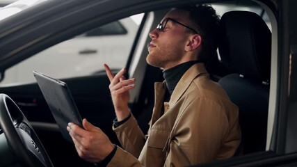 Stylish young man talking in the car on video via tablet