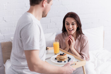 Happy woman looking at tasty pancakes and orange juice on tray near husband in bedroom