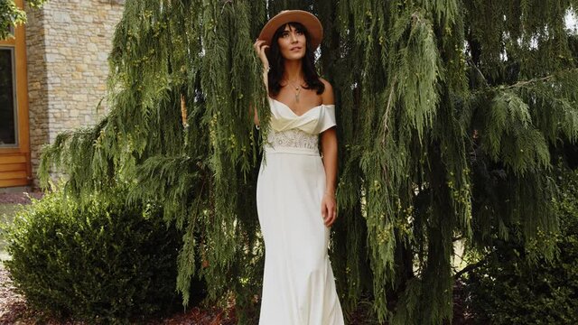 Woman in bridal gown with bohemian style fedora hat poses by pine tree, Slowmo