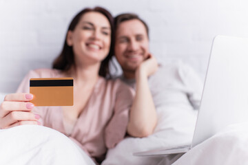 Credit card in hand of blurred woman near husband and laptop on bed